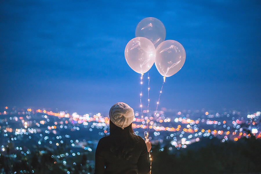 Person holding balloons looks out over an evening cityscape